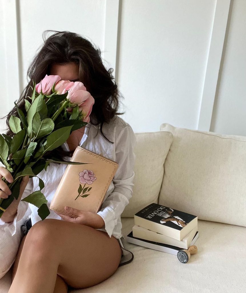 Halle sitting on a couch hiding her face with pink flowers; with a stack of books beside her