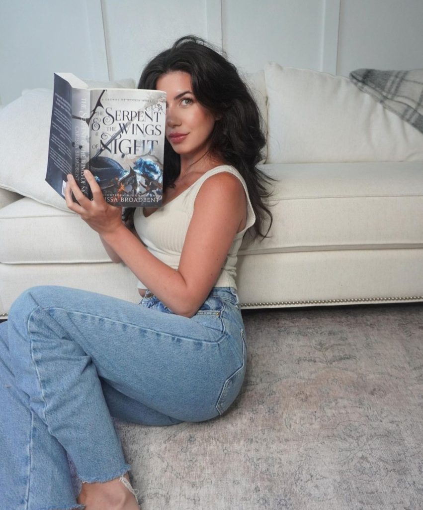 Halle sitting on the floor hiding half of her face with a copy of The Serpent and the Wings of Night by Carissa Broadbent