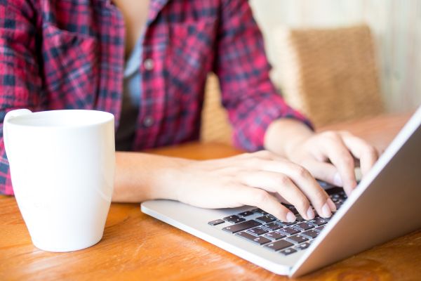 A woman in a flannel writing on a laptop with a white mug next to her.