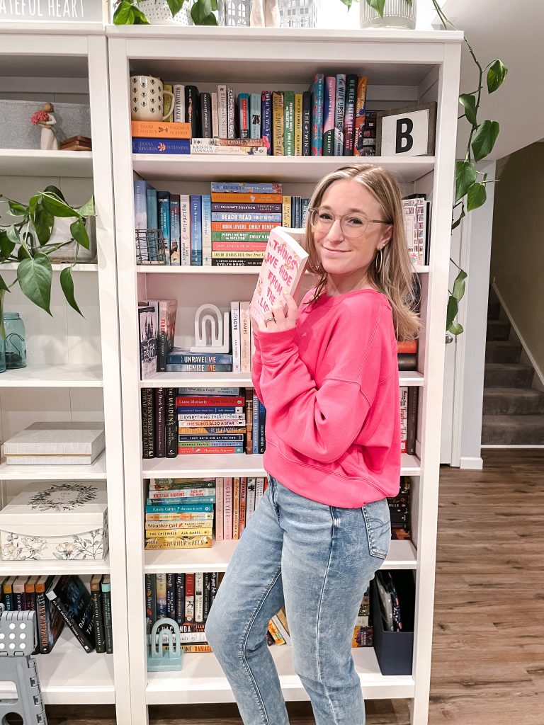 Jaclyn holding Lucy Score's Things We Hide From the Light book in front of her book shelves. 