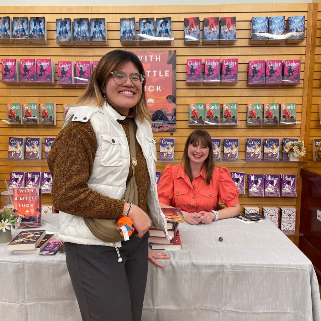 Picture of article writer with author Marissa Meyer signing her books, with background of Marissa Meyer's books. 