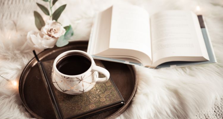 open book on a white blanket with a cup of coffee on a tray and flowers in the background