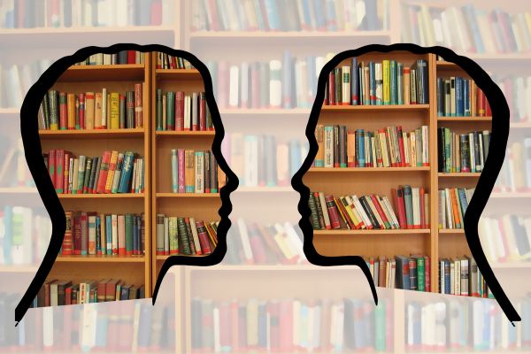 Two silhouettes of a people's heads face each other in conversation. Filled inside the outline of the faces are book shelves with many different, colorful books.