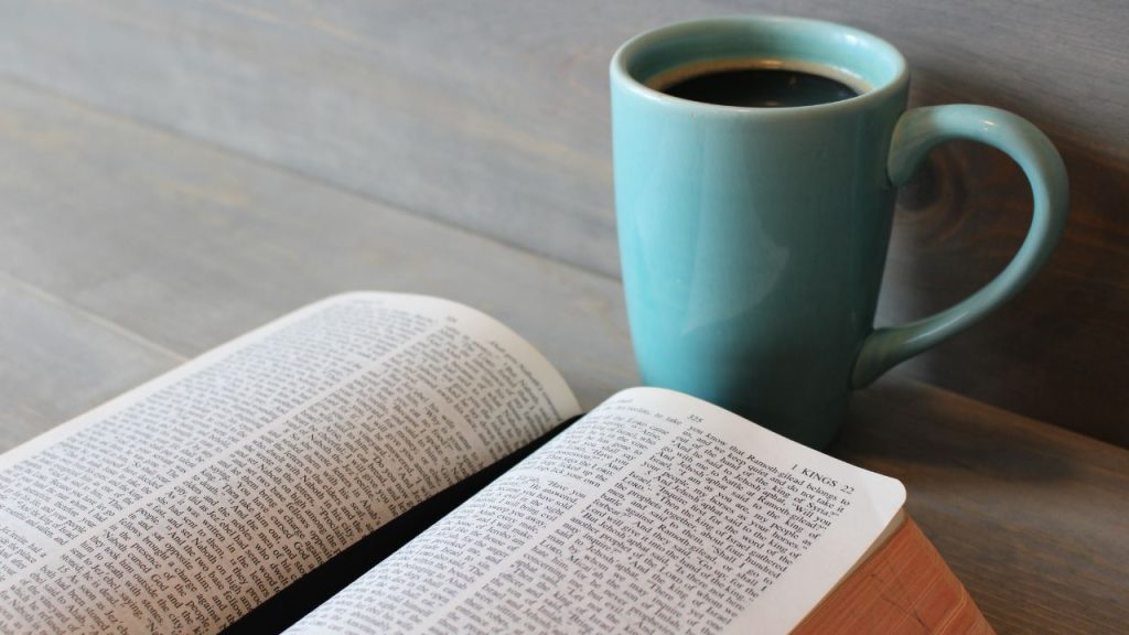 An open book with a black ribbon book mark sits on a gray, wooden table in front of a blur ceramic mug with black coffee.
