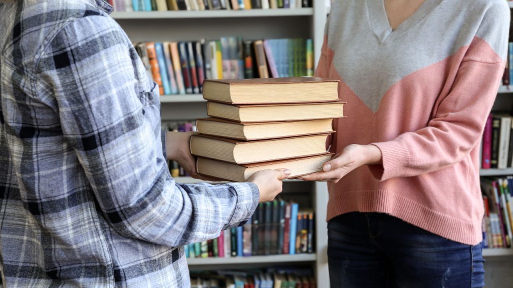 Two people exchange a stack of books in a library. On person on the right has a pink and gray chevron sweater, and the person on the left is in a gray flannel.