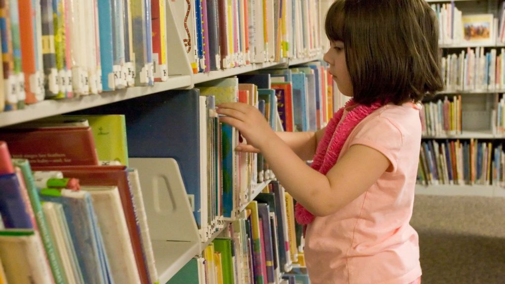 A young girl in a pink shirt and necklace rifles through picture books on a library shelf.
