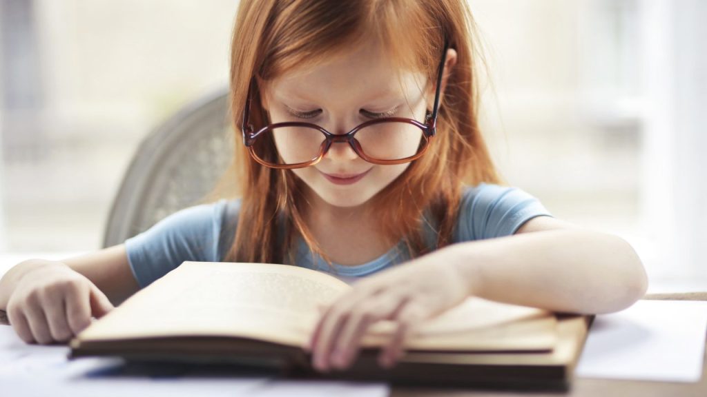 A young girl in glasses reading a book at a table.