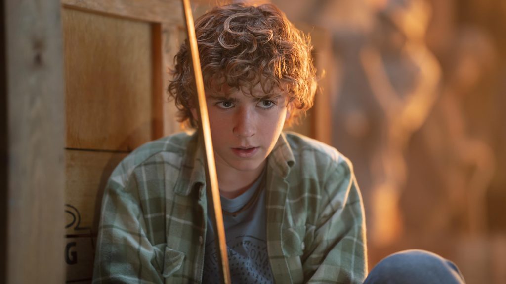 A boy with curly blonde hair wearing a green flannel and holding a gold sword. 
