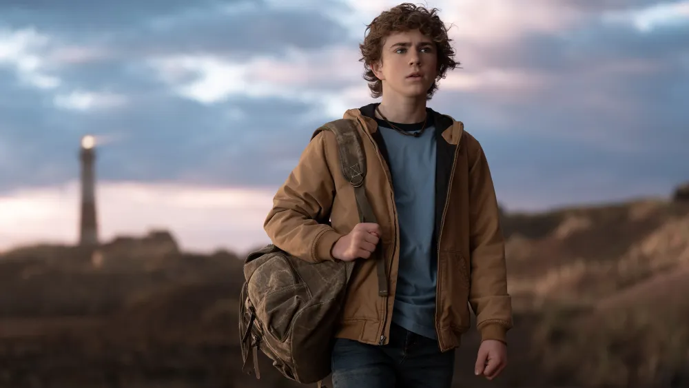 A boy with curly blonde hair wearing a khaki-colored jacket, a blue shirt, and has a backpack slung over one shoulder. 