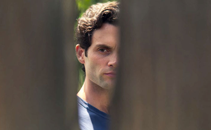 Brunette man in blue shirt looking between 2 fence posts with a serious expression