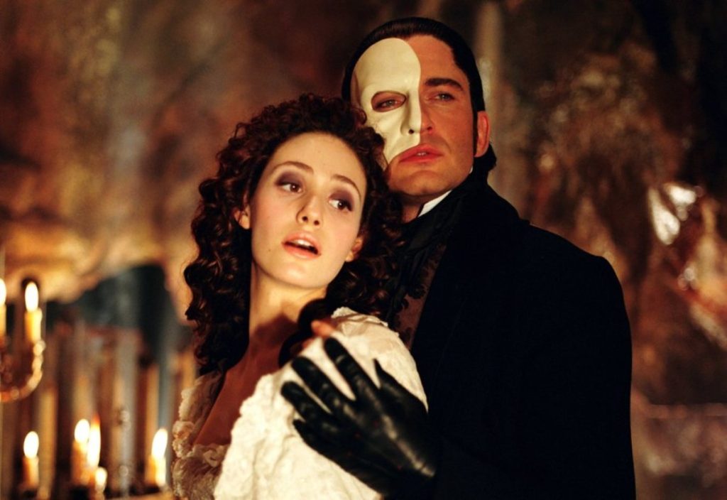 Brunette man dressed in black wearing a mask on half of his face holding a brunette woman in a white dress