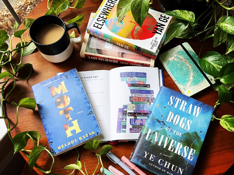 A collage of books are strewn on a dark brown wood table with plant vines around the edge. The books include a reading log, Straw Dogs of the Universe by Ye Chun, Moth by Melody Razak, and a celestial journal.