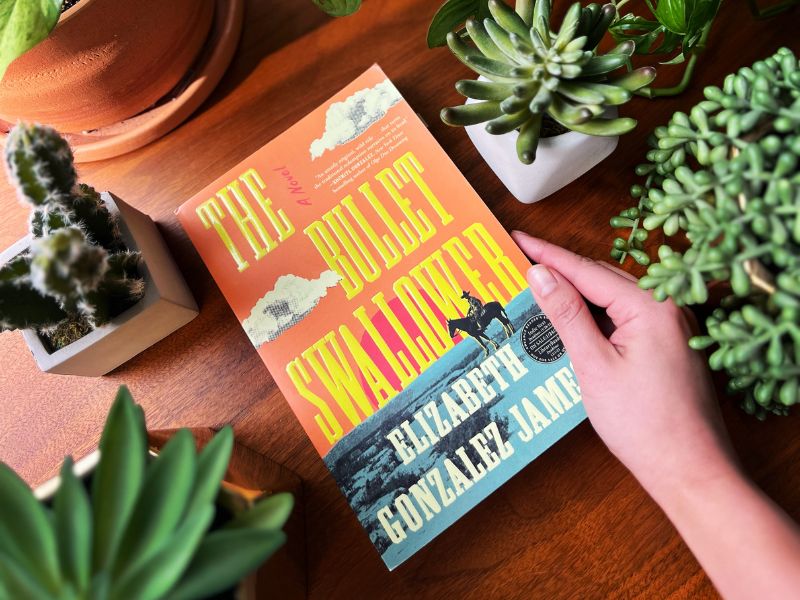A hand rests on the book The Bullet Swallower by Elizabeth Gonzalez James. On a dark brown table, multiple succulents and a cactus sit on the frame.