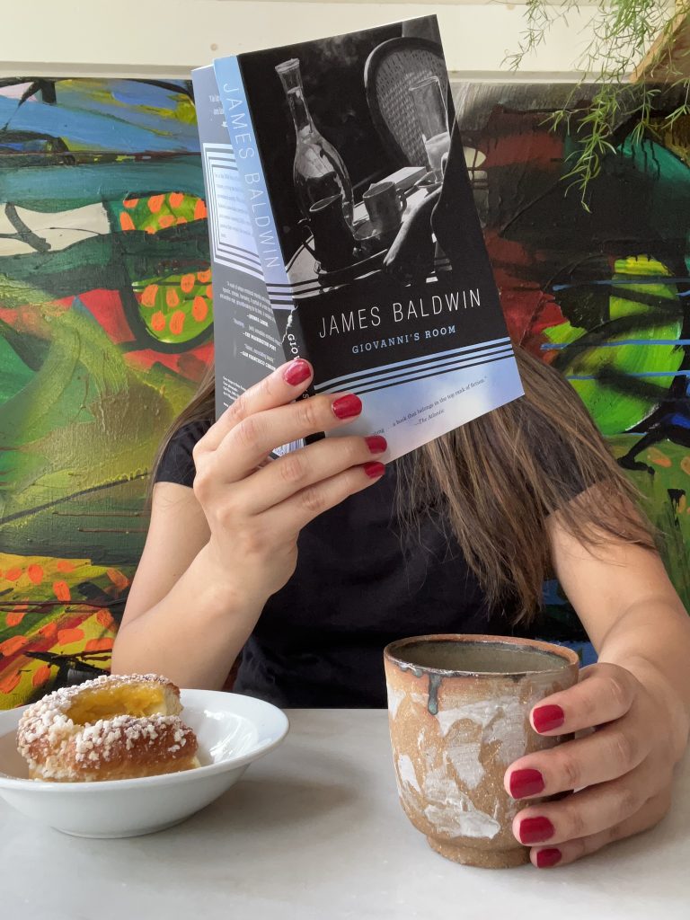 Bookstagrammer Kristin Lee holds Giovanni's Room by James Baldwin in front of her face. In her left hand is a mug, and on the white table in front of her is a plate with a pastry. The wall behind her has a mural and a handing plant on the right corner.