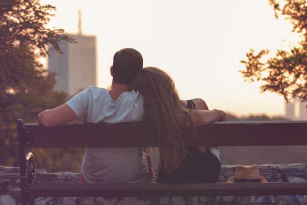 A young couple sitting on a bench facing a scenic view with their backs to the camera.