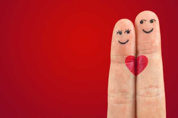 Two fingers with faces drawn on them on a red background looking at each other. A heart is painted below them to show they are in love.