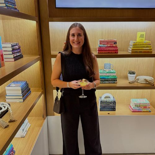 melissa holding a cocktail in a black jumpsuit in front of brown bookshelves with stacks of books color coordinated
