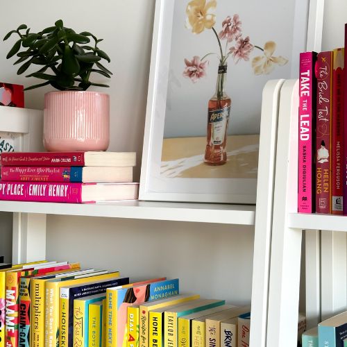 white bookshelf with a picture of flowers in a bottle of aperol spritz. also on the shelf is a succulent in a pink pot and pink and yellow books