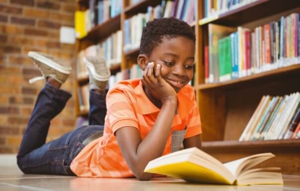 A young boy lying on the floor smiling while reading a book.