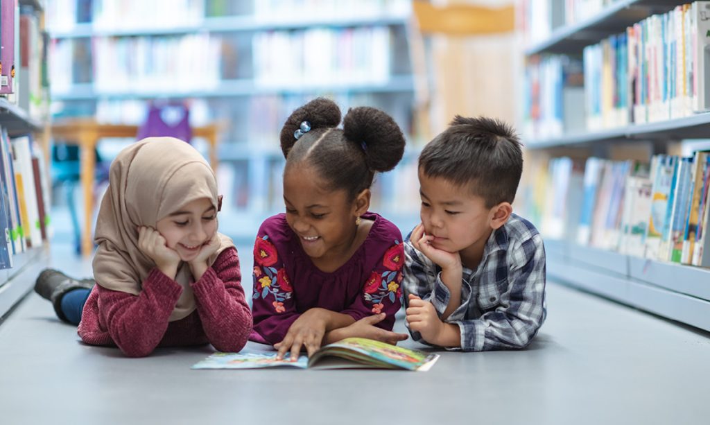 Three youngkids lying on the floor in a library reading.