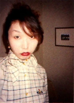Photograph of mangaka Ai Yazawa in a white plaid button-up shirt and red lipstick, posing in front of a wall with a single frame.