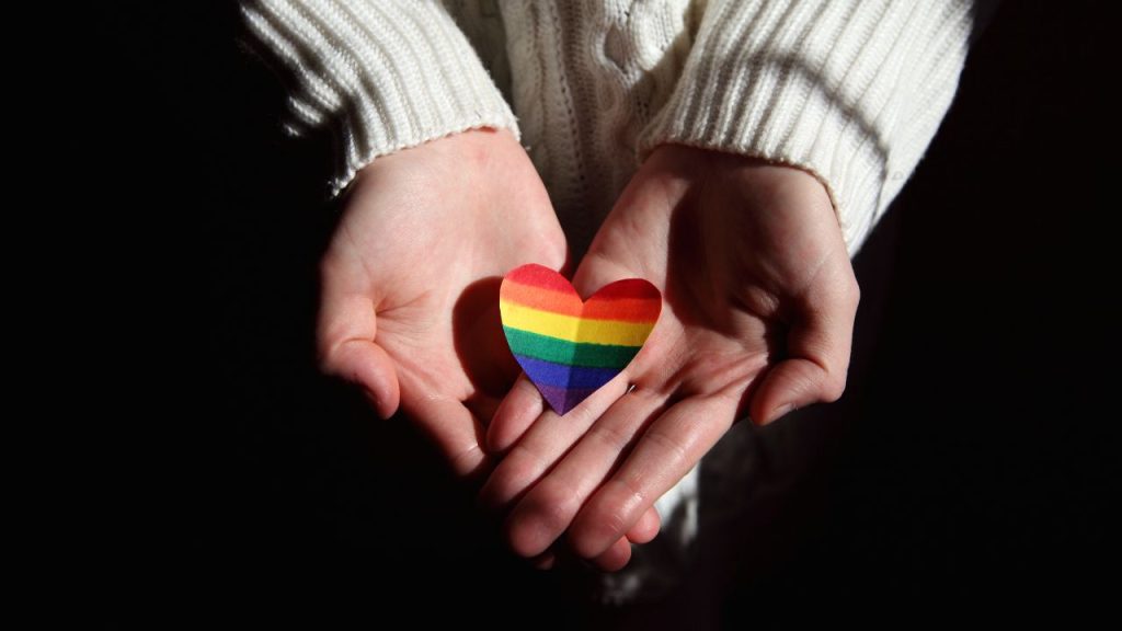 Two hands hold a paper heart with the pride flag colors drawn on it. The person holding out their hands is in a white cable knit sweater.