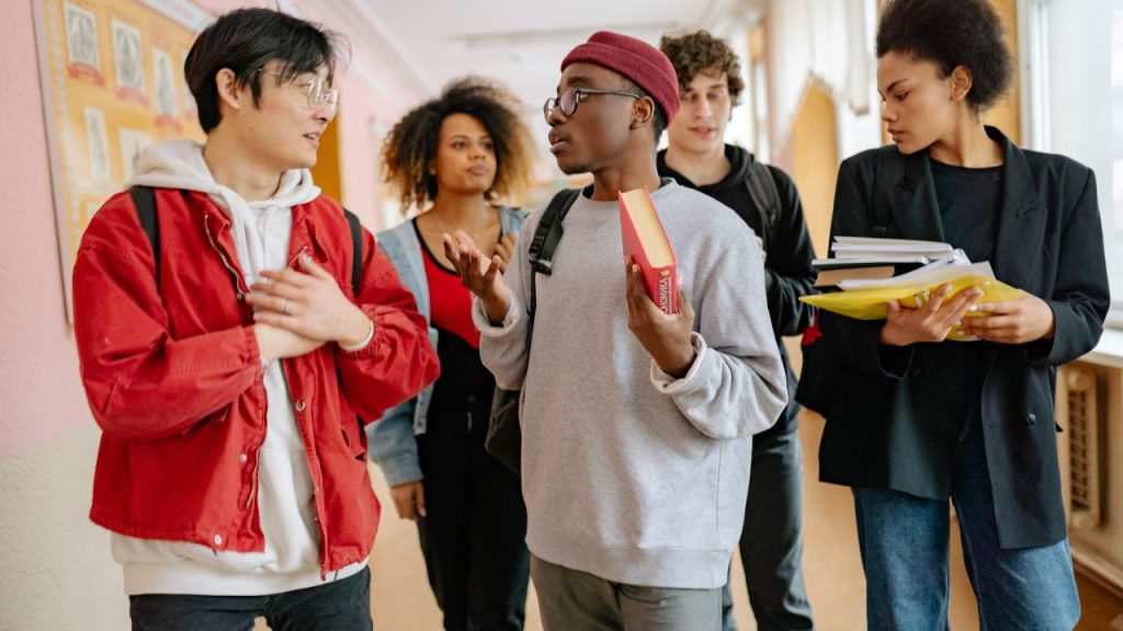 A diverse group of high school students walk and converse in a brightly lit school hallway. Some are holding books and others have book bags.