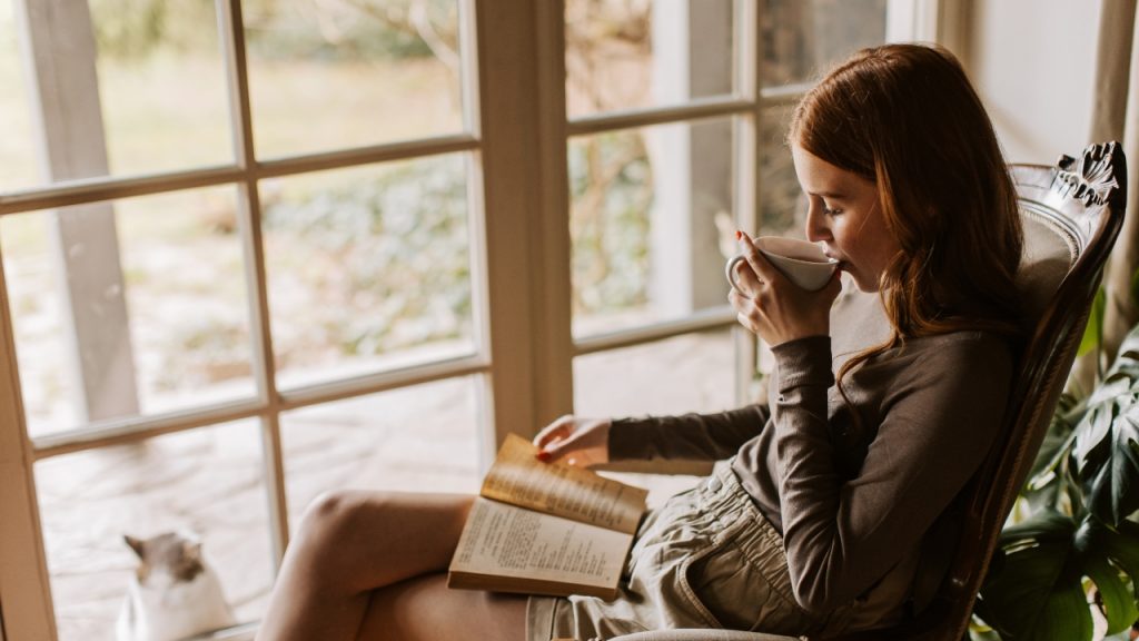 Woman reading a book on a chair while sipping a beverage, in front of glass panel doors that show a cat peering through from outside.