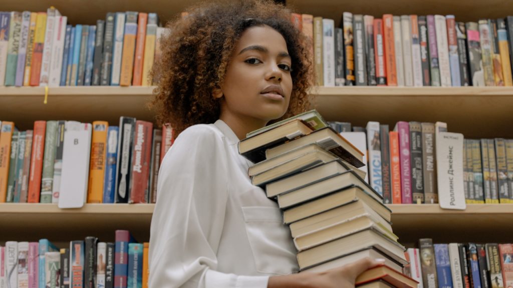 A young black woman in a white blouse looking into the camera while holding a stack of books, in front of library bookshelves.