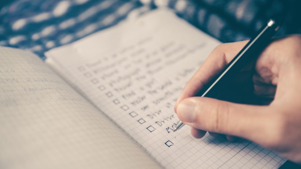 Close-up image of a hand holding a pen, writing a checkbox list on a graph grid page notebook.