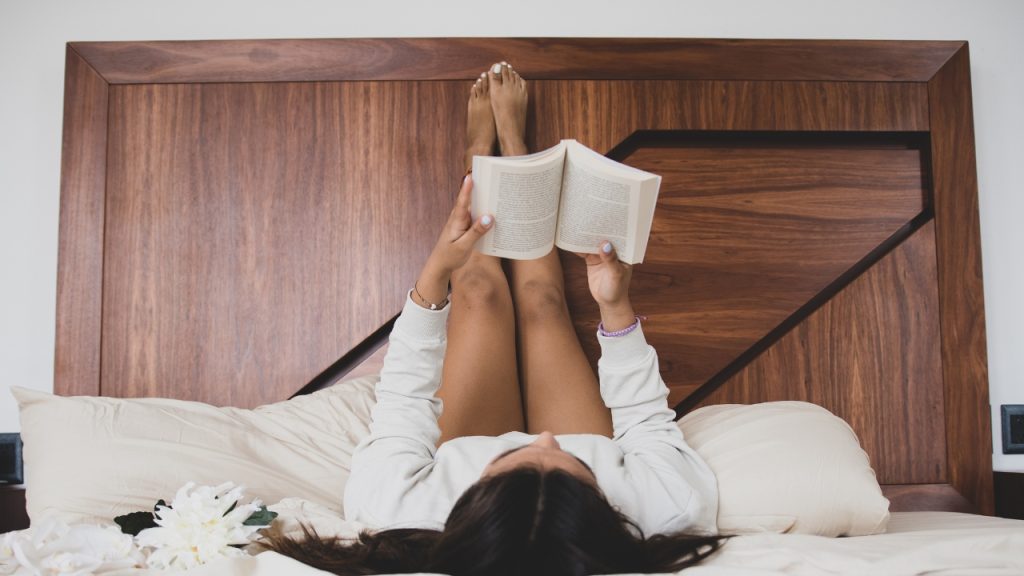 A young woman laying in bed reading, with her feet propped up against a wooden bedframe panel and a white flower lying next to her head.