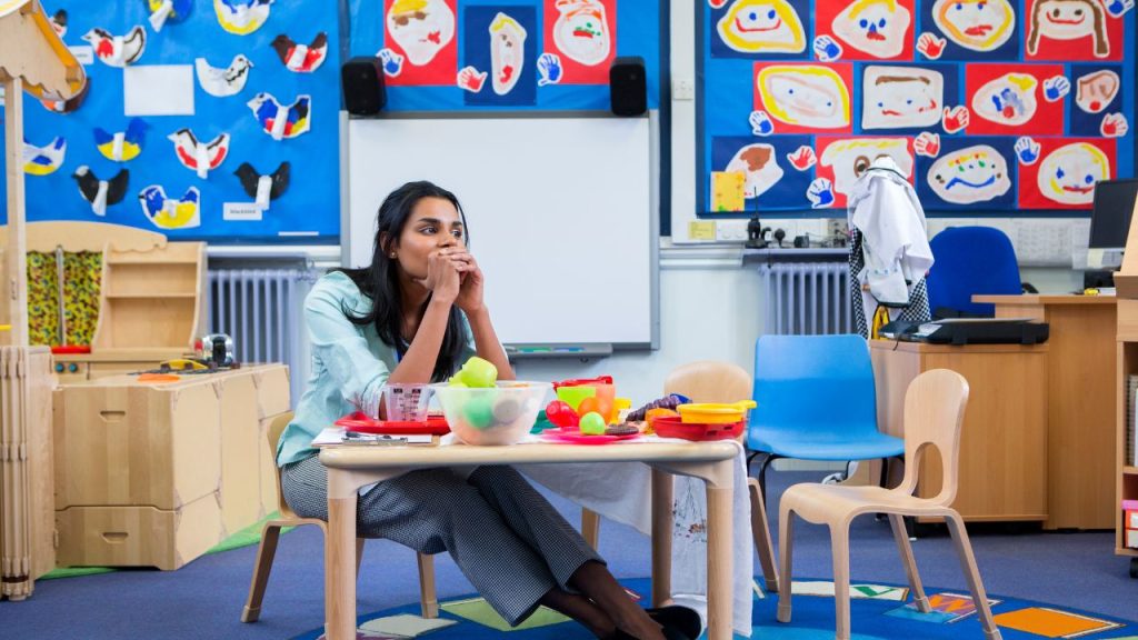 In a colorful elementary classroom, a teacher in a blue button-up and grey slacks stares distantly in frustration with her head in her hands. She sits at a wooden table made for children with colorful craft tools in containers.