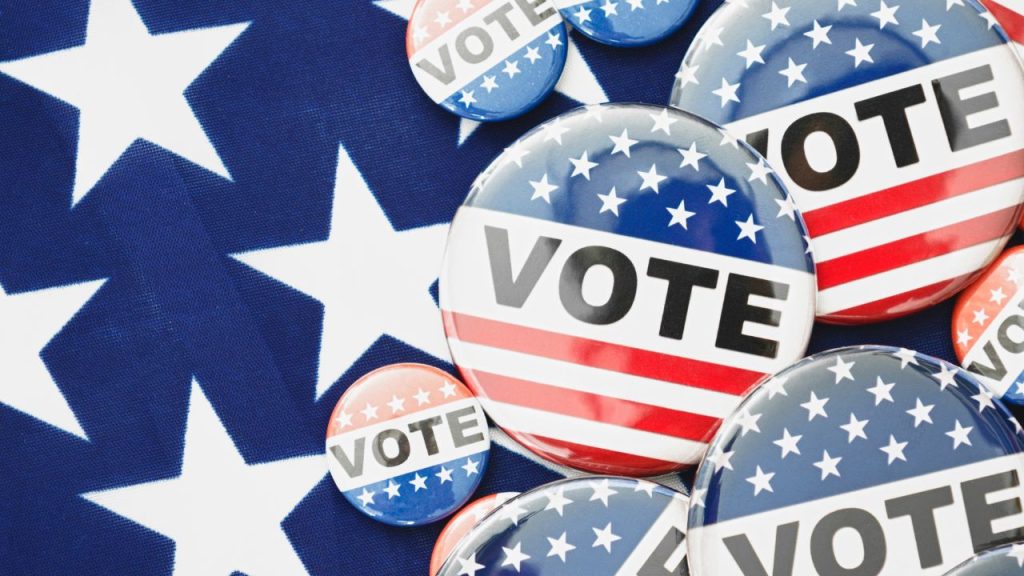American flag pins with the word "Vote" in black are clustered on a cloth with an American Flag star print.