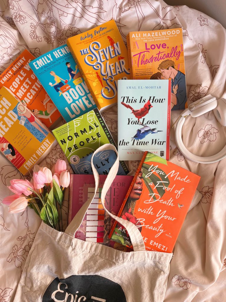 A flat lay of assorted books against a bedsheet. A pair of white headphones are on the right side with a bouquet of pink flowers to the left.
