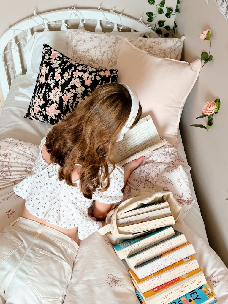 A woman in a white shirt and pants lies on her bed on her stomach reading a book. She's wearing a white pair of headphones with a totebag full of books next to her.