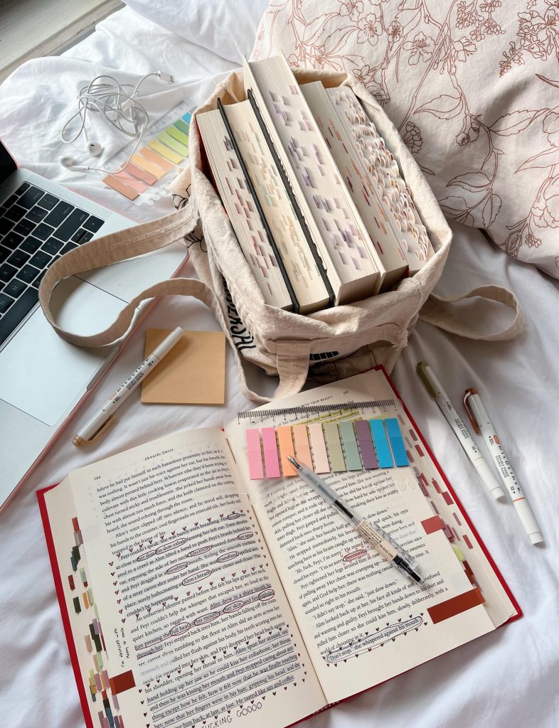 An open and annotated book is open towards the bottom of the image surrounded by pens and highlighters. A laptop lies to the left side and a tote bag of books sits in the center.