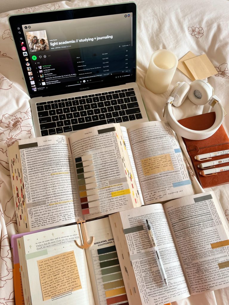 Four open and annotated books take up the bottom of the image with an open laptop at the top. A white candle, white headphones, and a red notebook with highlighters rest to the right side.