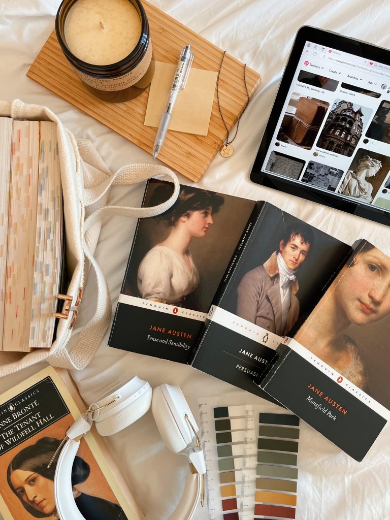 Flatlay against a white bedsheet with three Jane Austen books to the center right, an Ipad in the top right, and a brown board in the center left. A tote bag with annotated books is to the center left.