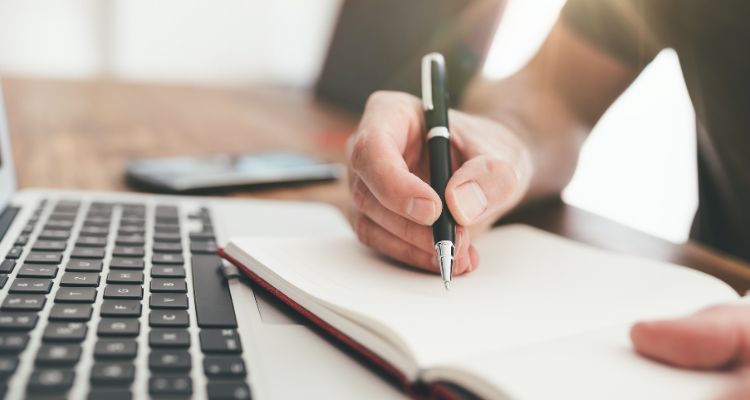 Man writing notes in a notebook before an open laptop