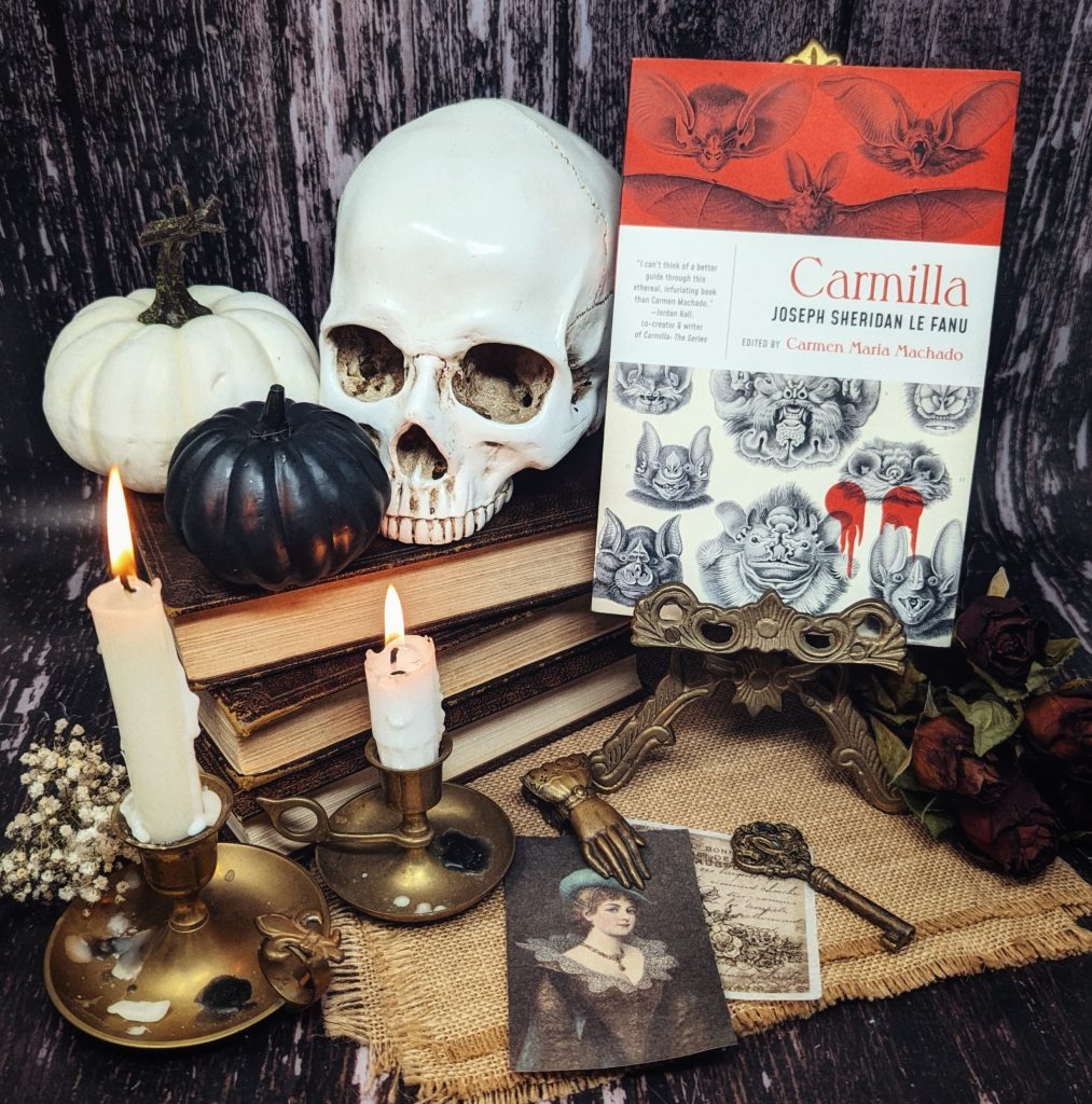 The book Carmilla by Joseph Sheridan Le Fanu is held by an ornate gold stand. A stack of old books behind it holds a skull decoration and a white and black pumpkin. Two tall, white candles burn on gold plates with white flowers at the base. Vintage photos of a woman and a letter sit in front of the book with a key and hand-shaped charm on top. Behind the book are a couple of dried roses.