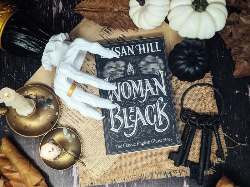 The book Woman in Black by Susan Hill is positioned on a black wooden table. Beneath is beige cloth and a yellowing newspaper page. Surrounding them are white and black pumpkins, a set of vintage keys, two tall, lit candles on gold plates, autumn leaves, and a white skeleton hand with a gold ring on one of its fingers.