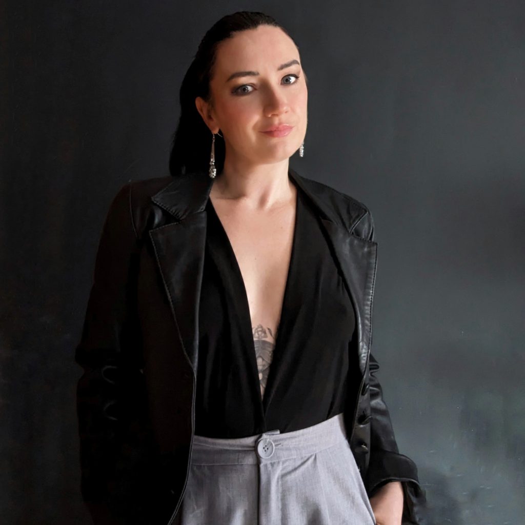 Photo portrait of Bookstagrammer Marie McWilliams. She poses in front of a gray background, in a black leather jacket and blouse, with her hand in the pocket of her light gray trousers.