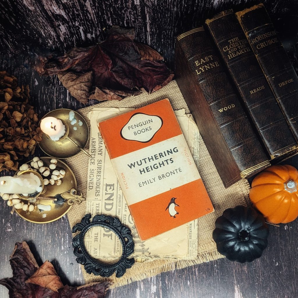 Wuthering Heights by Emily Bronte is positioned on top of a piece of beige cloth and a yellowing newspaper. To the right are three brown leather books of the same size and orange and black pumpkins. To the left are two tall white candles, lit and dripping on gold plates. A mirror frame and dried autumnal flowers and leaves decorate the available space.