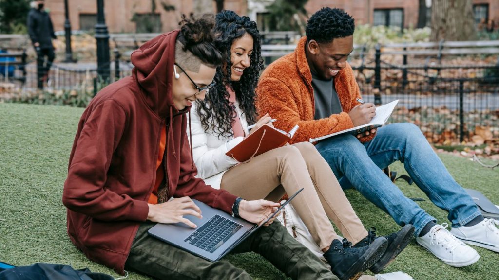 Three happy university students sit together on a lawn in a city. The first man in the line of three types on a laptop in a red hoodie. The woman next to him writes in a leather journal and the other man next to her writes in a notebook. The woman is wearing a white puffer jacket and khakis, and the last man is in an orange jacket and jeans.
