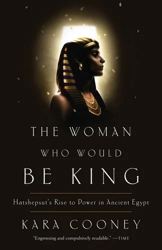 A model dressed as Hatshepsut stands in the background of the image against a black background and a ring of gold light surrounding them.