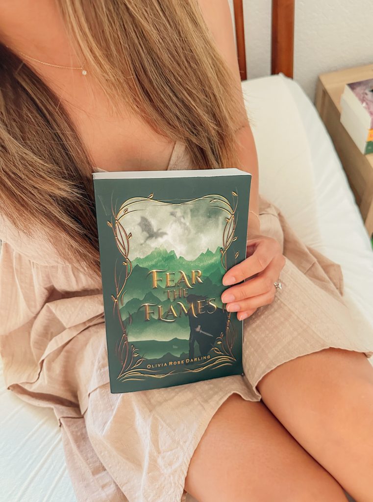 Bookstagrammer Jessica @jessabibliophile holds Fear the Flames by Olivia Rose Darling. She is in a beige linen dress and sitting on a mattress. On the nightstand is a colorful stack of three books.