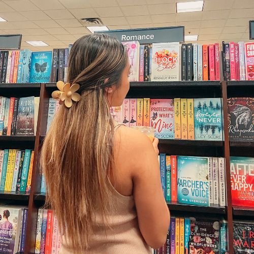 Bookstagrammer Jessica @jessabibliophile stands in front of the romance bookshelves in a Barnes and Noble. Her long brown hair is half up, held together with an orange flower clip.