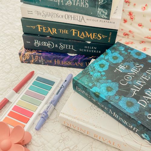 A bookish collage features a stack of fantasy novels in the background and two other fantasy novels laid out in front of the stack. A purple and pink pen lay on both sides of the annotation tabs, which are in rainbow colors. A pink flower hair clip is beside the tabs, and everything is resting on floral printed and embroidered sheets.