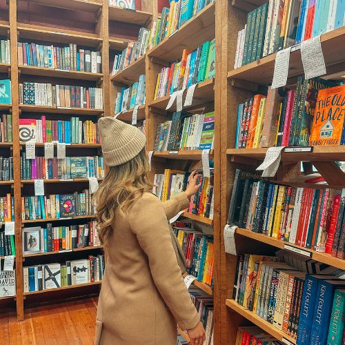 Bookstagrammer Jessica @jessabibliophile standing in front of bookstore shelves. She is in a beige winter coat and a lighter beige beanie. She reaches out to grab a book off the shelf.