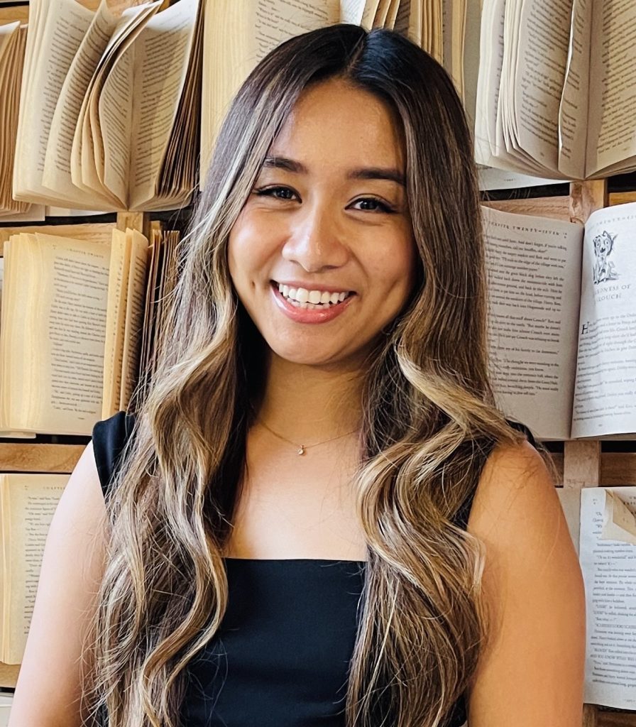 Portrait of Bookstagrammer Jessica @jessabibliophile. She has long wavy brown hair, a black tank top, and is standing in front of a wall with open books.